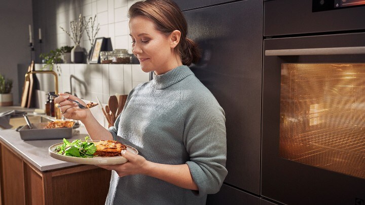 En kvinna står lutandes i köket och äter en nylagad lasagne.